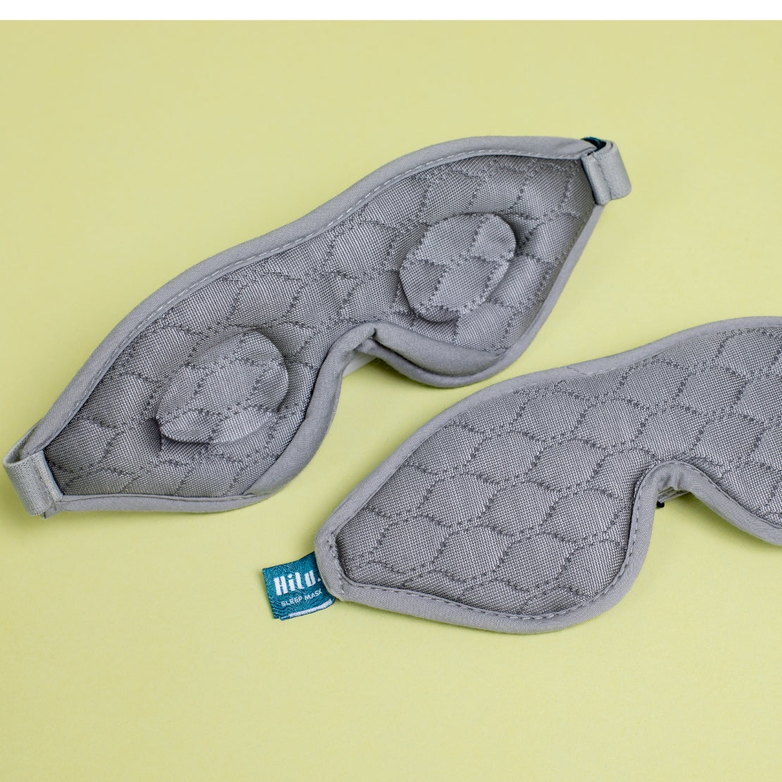 Two gray sleeping masks with textured quilted patterns, placed on a light yellow background. One mask is flipped to show the inner soft cushioning.