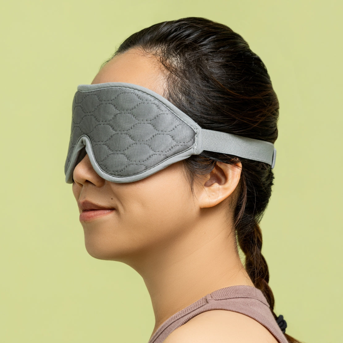 A woman wearing a gray textured sleeping mask that covers her eyes, with an adjustable strap around her head. The mask appears padded for comfort