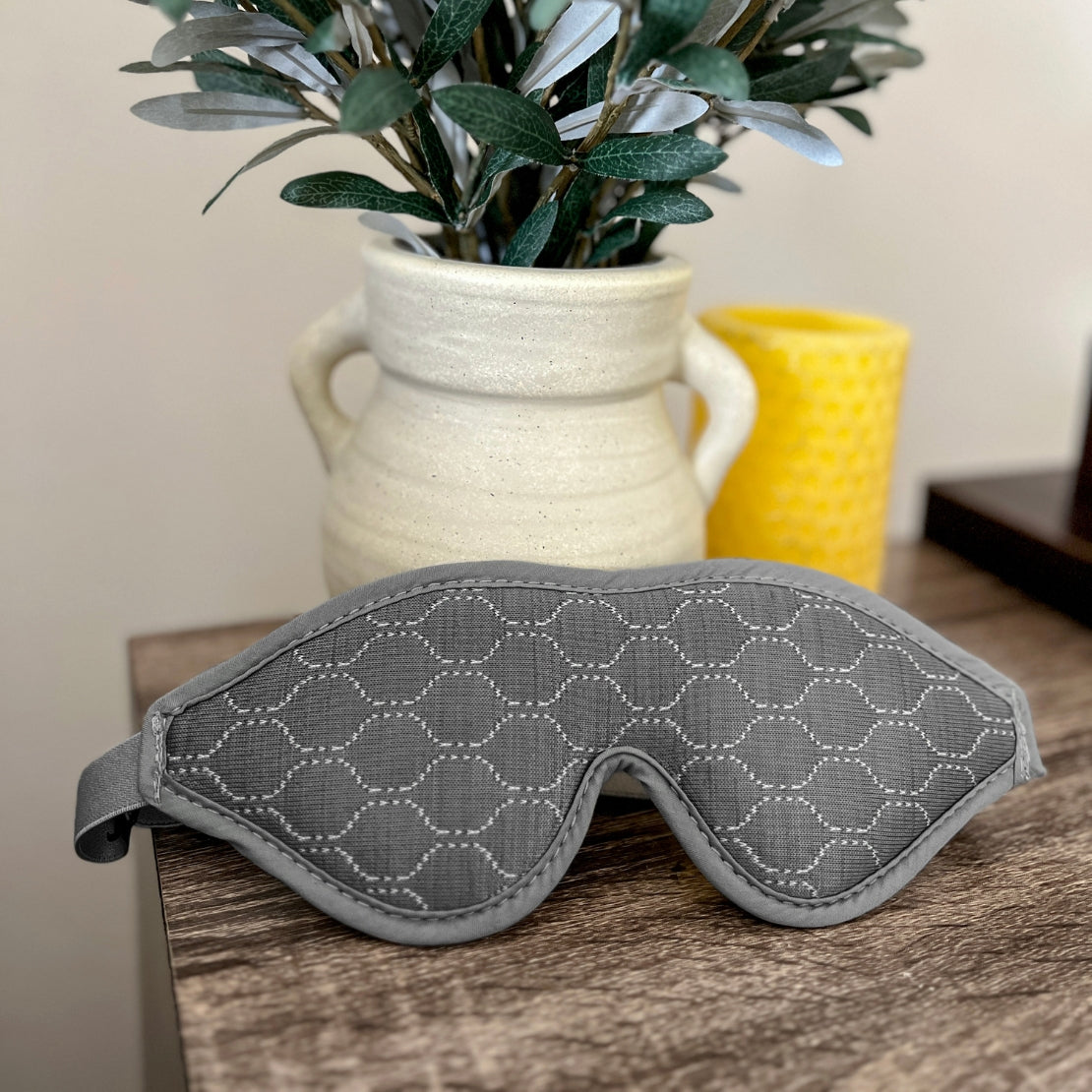A close-up view of a gray sleeping mask with a hexagonal pattern, placed on a wooden surface. In the background, a ceramic vase holds green foliage next to a bright yellow cup