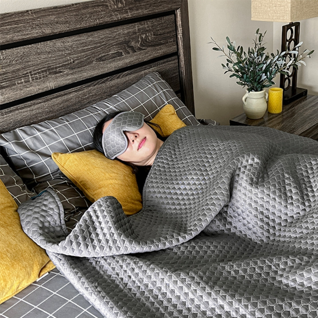 A person resting on a bed with a gray HILU Sleeping Mask covering their eyes, surrounded by yellow and gray pillows, with a wooden headboard and decorative plants in the background.