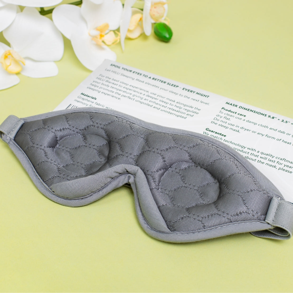 Grey HILU sleeping mask with quilted pattern placed on a yellow background alongside white orchid flowers.