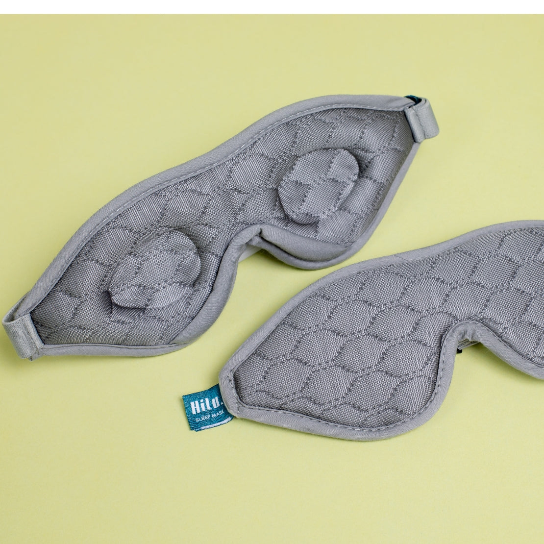 Two gray sleeping masks with textured quilted patterns, placed on a light yellow background. One mask is flipped to show the inner soft cushioning. 