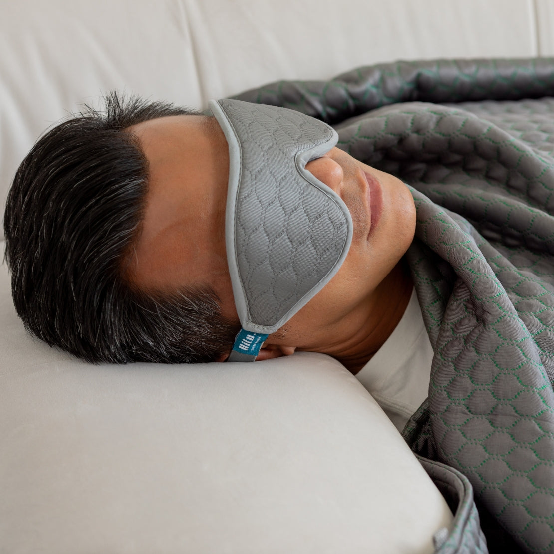 A person lying down comfortably with a textured grey HILU sleeping mask covering their eyes, complemented by a quilted green blanket.