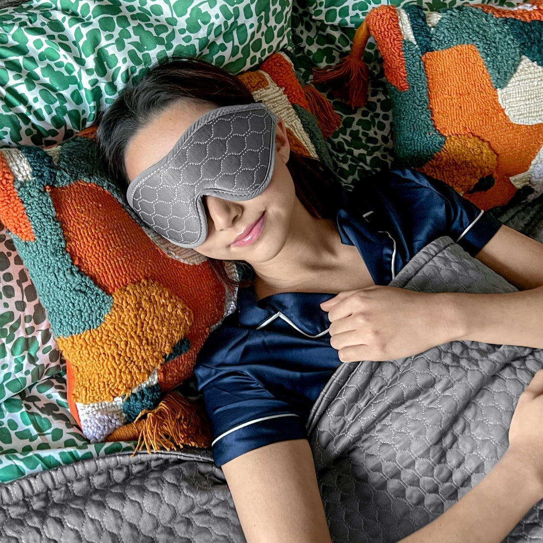 A person comfortably resting on a bed with a gray patterned HILU sleeping mask covering their eyes, surrounded by colorful and textured pillows.