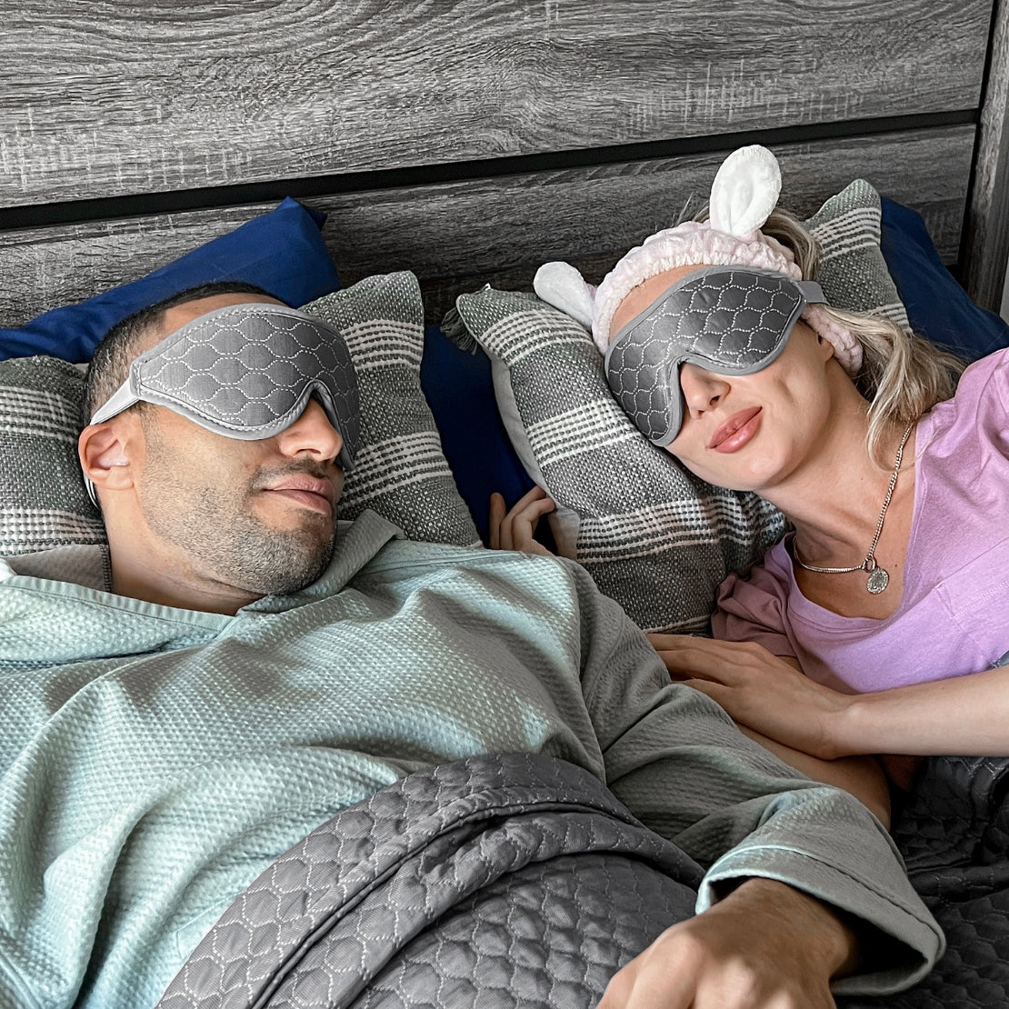 Two individuals lying side by side on a bed, each wearing unique sleeping masks. The man on the left wears a gray patterned mask, while the woman on the right sports a mask with bunny ears. They are surrounded by comfy pillows and blankets, emphasizing a peaceful sleep environment