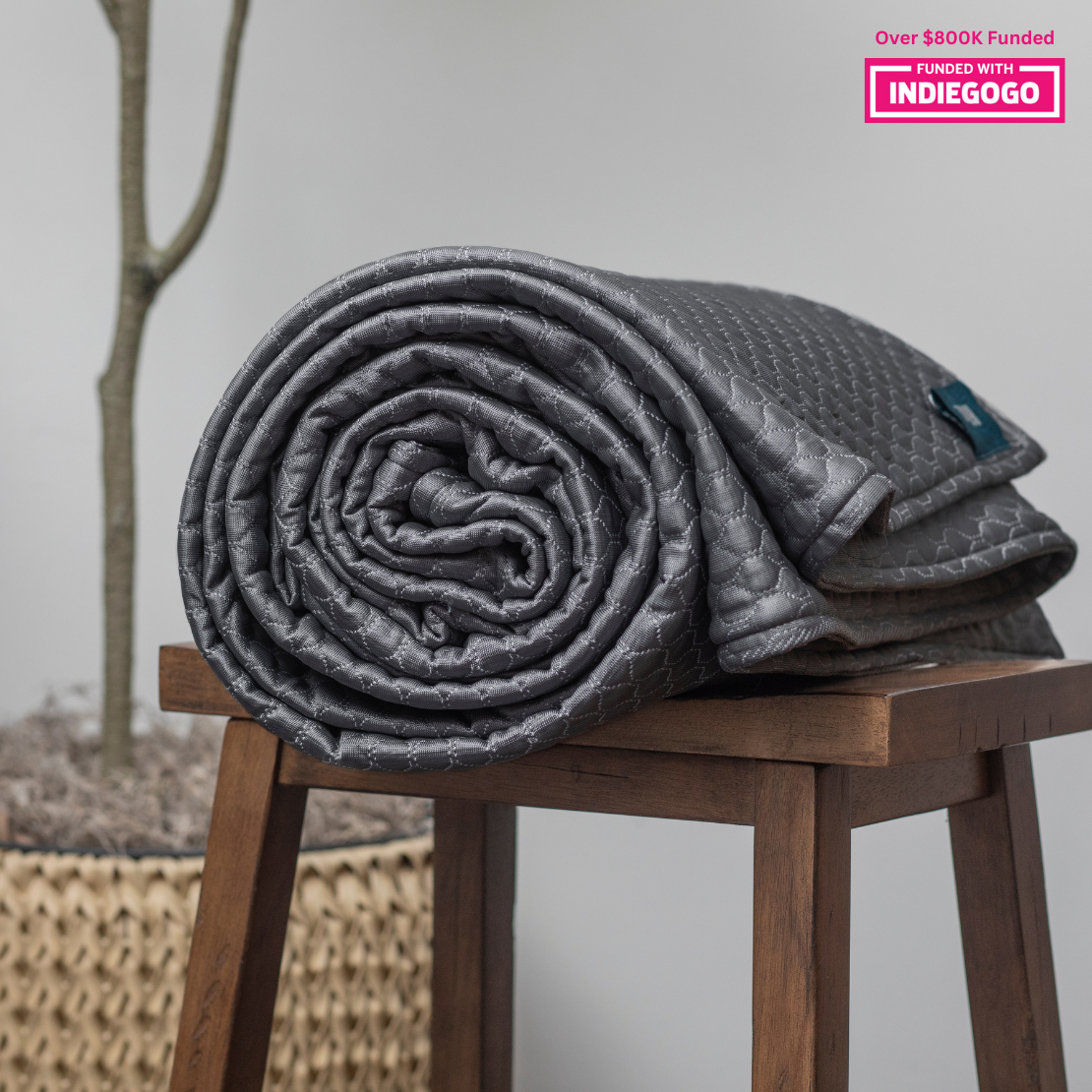 A neatly rolled gray quilted blanket with a diamond pattern rests atop a wooden stool. The blanket displays intricate stitching and has a small label at one end. In the background, there's a blurred view of a tall thin tree and a woven basket.
