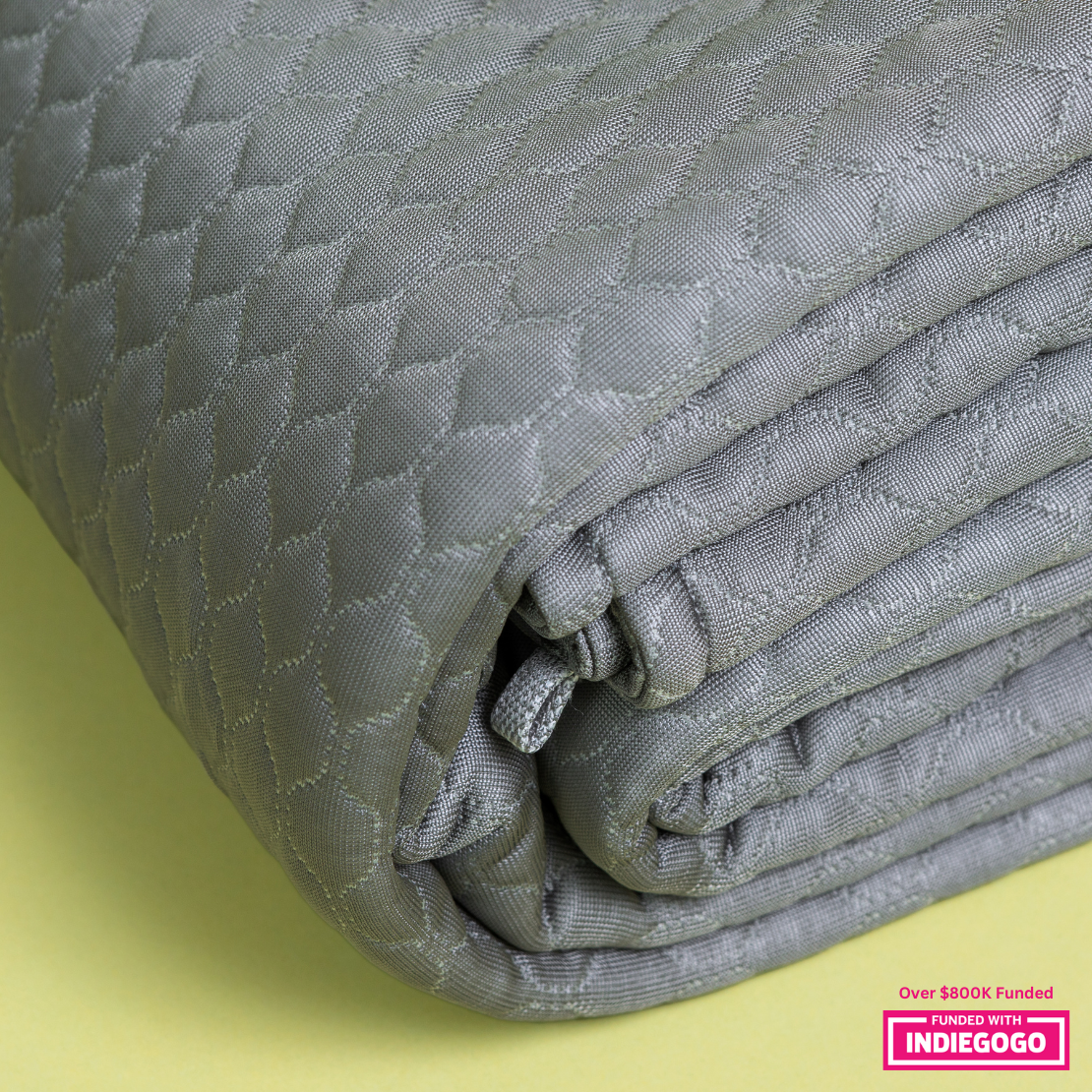 Close-up view of a silver-gray quilted blanket with a textured diamond pattern, neatly folded to showcase its intricate design against a pale yellow background.