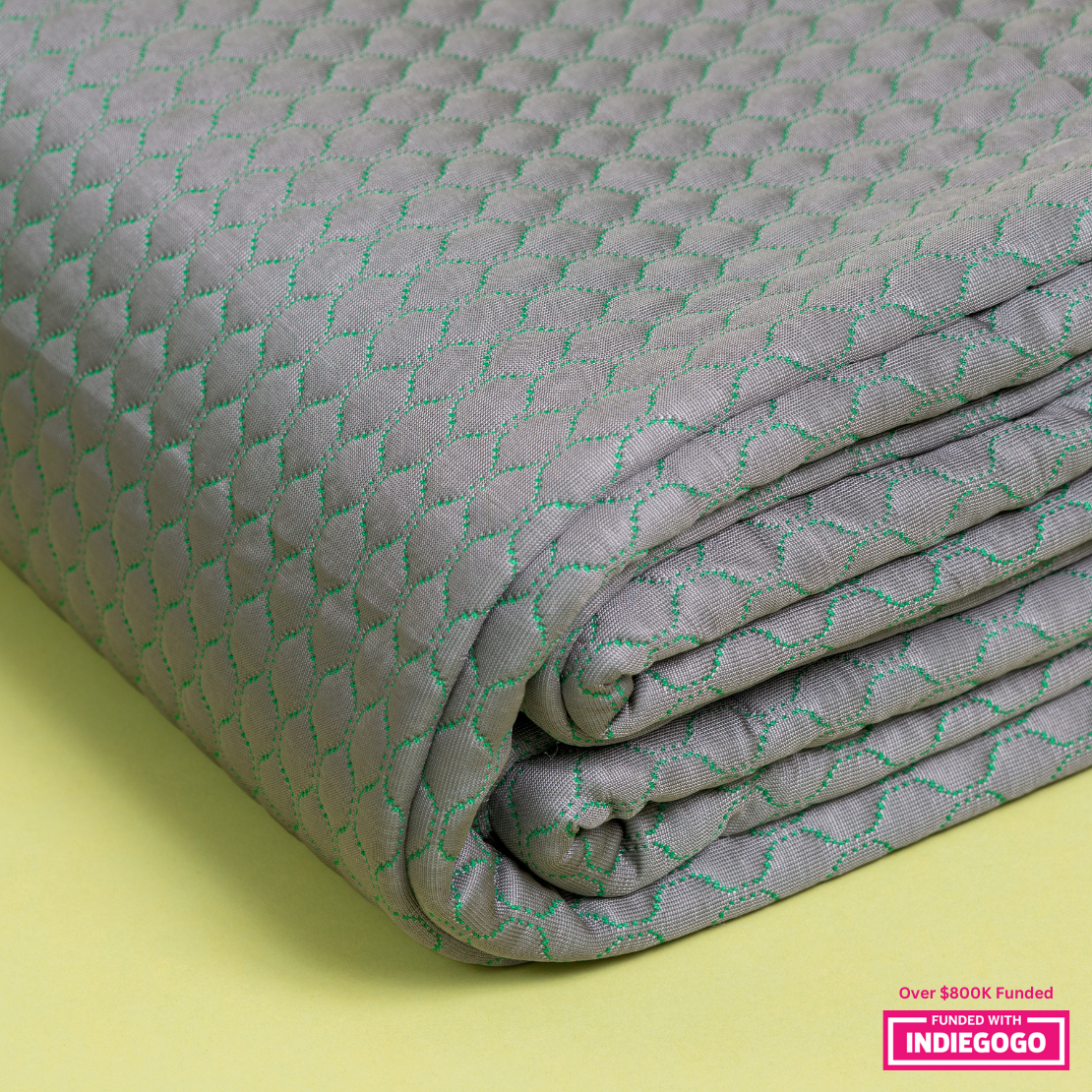 A close-up view of a textured, light gray blanket with a unique diamond pattern stitched in green thread. The blanket is neatly folded, showcasing its plush thickness against a soft yellow background.