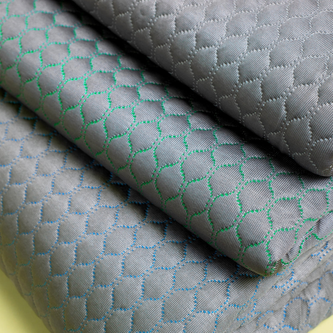 Close-up view of a textured blanket with intricate diamond-shaped patterns in shades of gray, highlighted by subtle green and blue stitching.