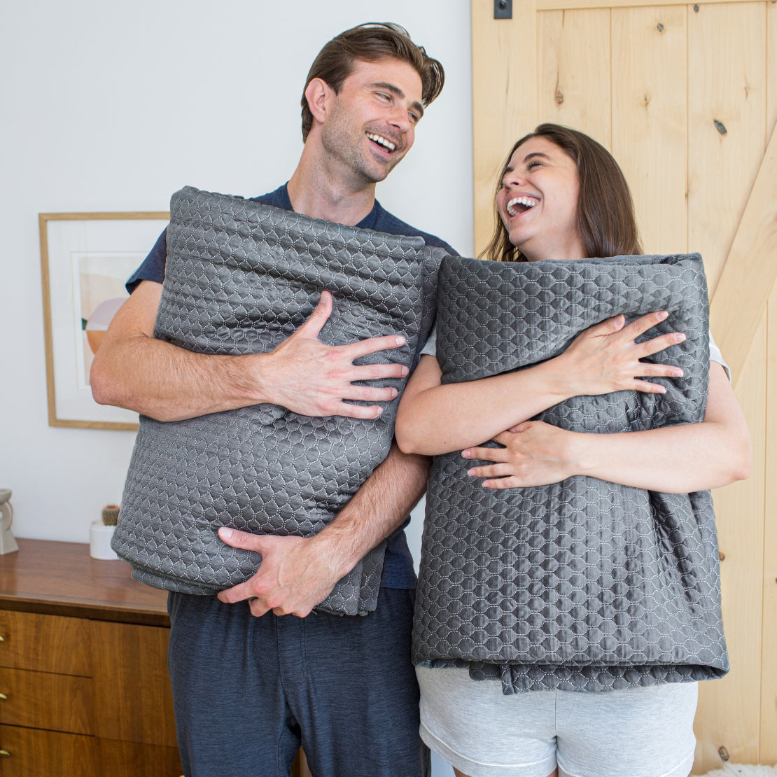 Two individuals joyfully holding large gray temperature-regulating blankets. The man on the left smiles and looks towards the woman, who is laughing. Both stand in a well-lit room with wooden features and minimalistic decor.
